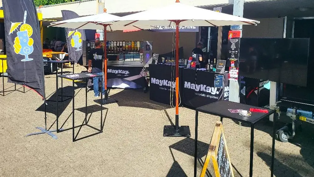 MayKay Cocktail-Showbar an einem Anlass in der Region Basel: Cocktailmaschine mit Flaschenwand unter Sonnenschirmen, Stehtische und Theke mit MayKay-Logo